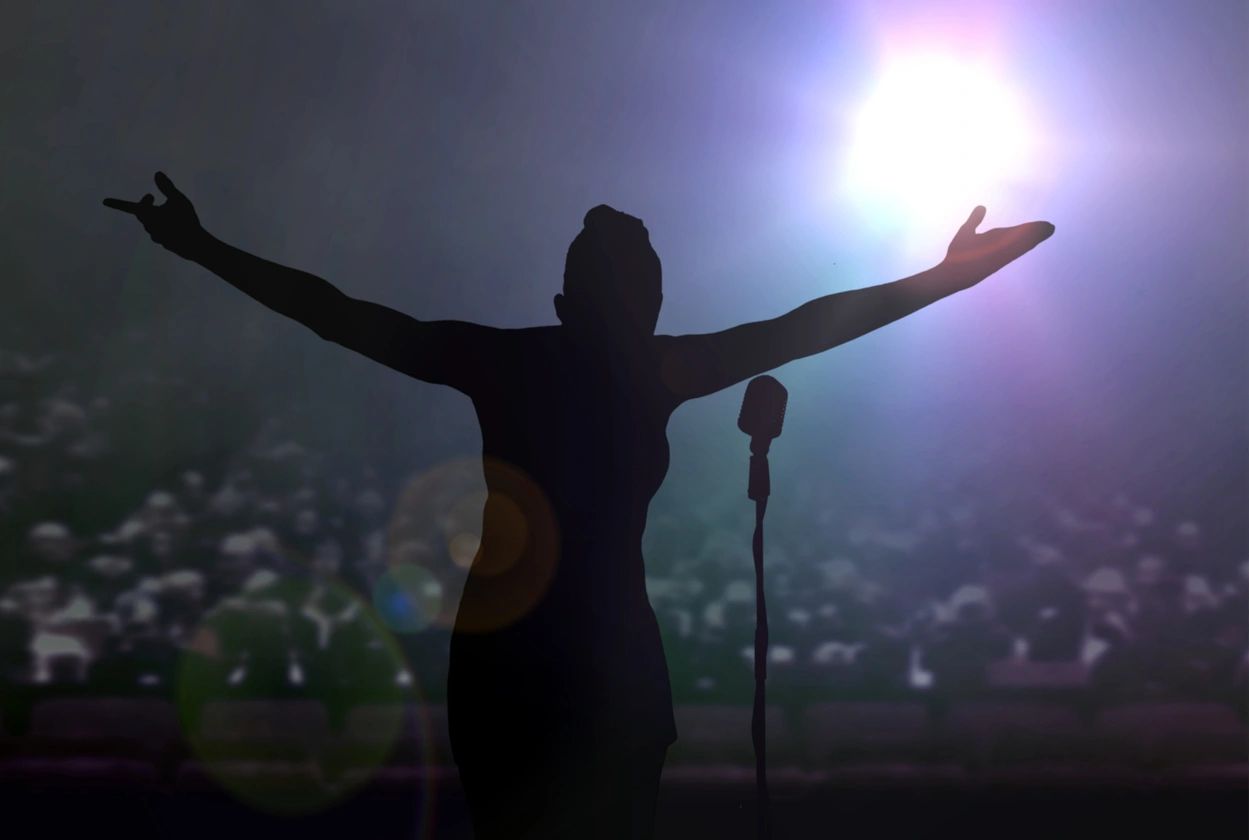 Silhouette of a performer with arms outstretched in front of a crowd and stage lights.
