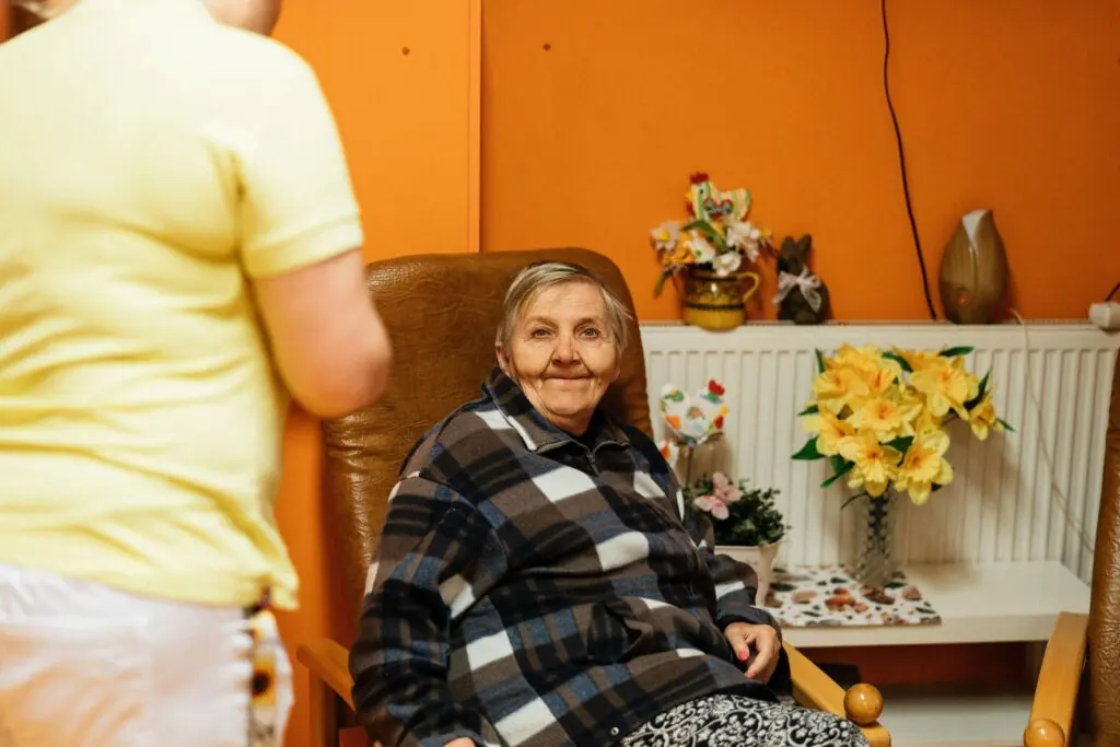 An older woman seated in an armchair with another person standing nearby.